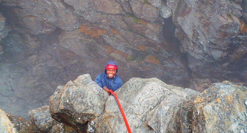 A person wearing safety gear is secured by ropes as they look up at the camera and smile while rock climbing.